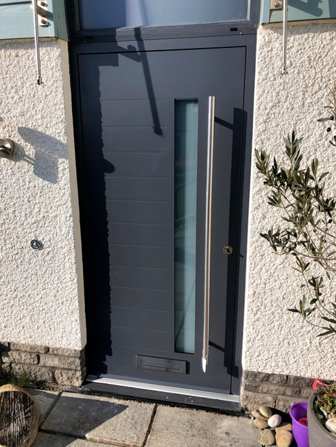A modern aluminium entrance door with a brushed steel handle and a triple glazed laminated glass insert, set in a dark brick wall with a subtle shadow on the wall behind.